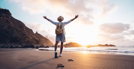 Mann in Rückansicht mit ausgebreiteten Armen läuft auf einem Sandstrand Richtung mehr und Sonnenuntergang