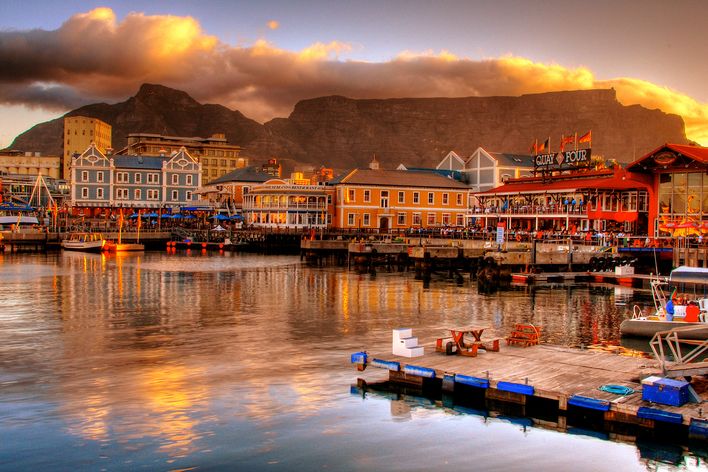 Kapstadts V&A Waterfront leuchtet bei Sonnenuntergang vor dem wolkenverhangenen Tafelberg