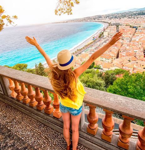 Frau mit Hut und ausgestreckten Armen blickt von einer Aussichtsterrasse über die Küste und Stadt von Nizza