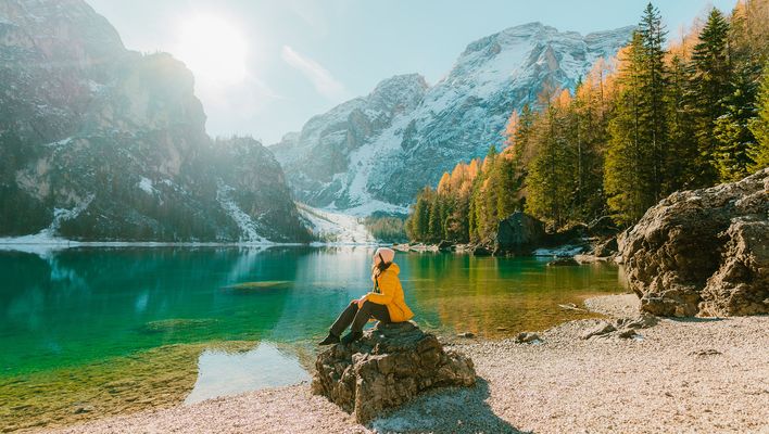 Frau sitzt auf einem Felsen vor einem See umgeben von sonnenbestrahlten Bergen und Bäumen in Südtirol