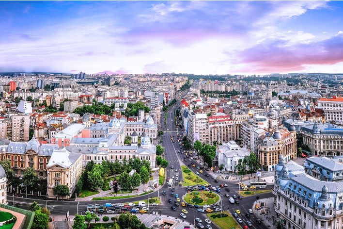 Über Bukarest erstrecken sich dicht bebaute Viertel und eine breite Allee unter violettem Abendhimmel.