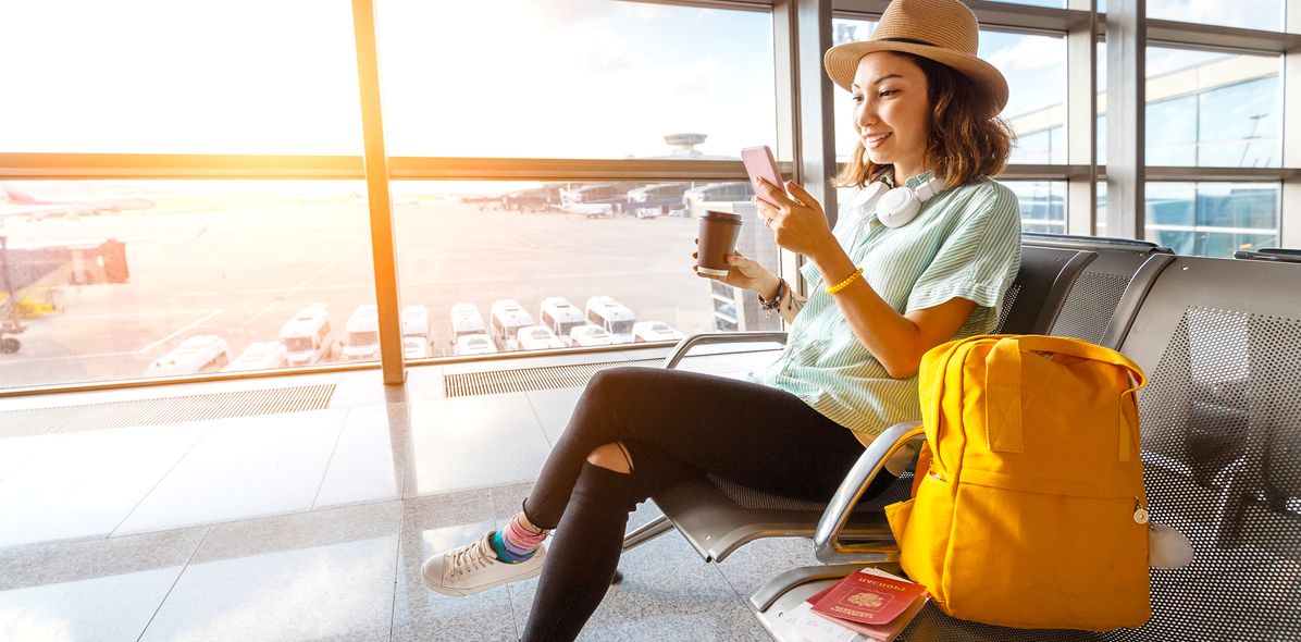 Frau mit Hut sitzt in einem Flughafen-Terminal am Fenster zum Rollfeld mit Handy und Kaffeebecher neben einem gelben Rucksack
