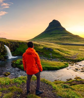 Ein Mann in einer roten Jacke steht auf einem Felsen und schaut auf den Kirkjufell Wasserfall in Island