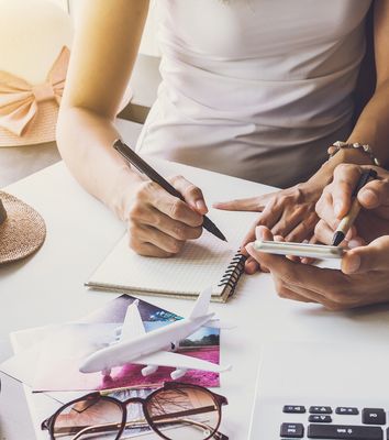 Hände eines Paars am Schreibtisch über Laptop, Karten und Globus, Urlaubsplanung am Tisch