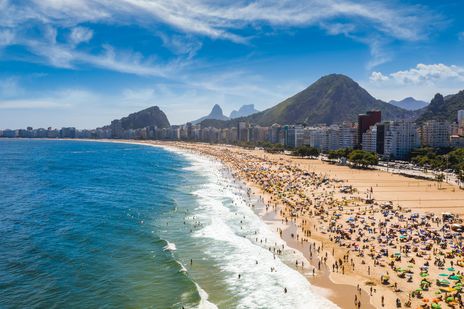 Die Küste von Rio de Janeiro zeigt einen belebten Sandstrand, türkisblaues Meer und Berge unter leicht bewölktem Himmel.