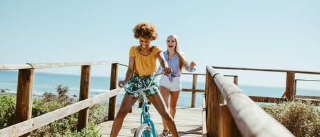 Frau, gefolgt von weiterer Frau, fährt mit Fahrrad über Holzsteg am Strand bei sonnigem Wetter