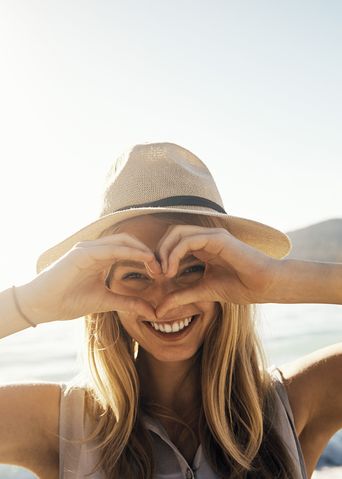 Frau mit Sommerhut am Strand formt mit den Händen ein Herz und schaut hindurch