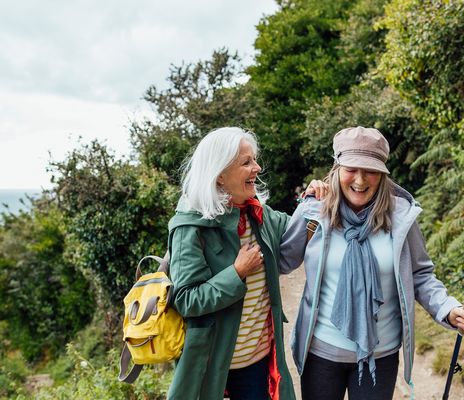 Zwei Freundinnen wandern lachend mit Rucksäcken auf sonnigem Waldweg