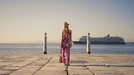 Frau mit einem bunten langen Kleid steht an einer Promenade und schaut auf ein Kreuzfahrtschiff
