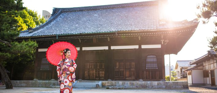 Frau im Kimono mit rotem Schirm vor traditionellem Tempel in Japan bei Sonnenlicht