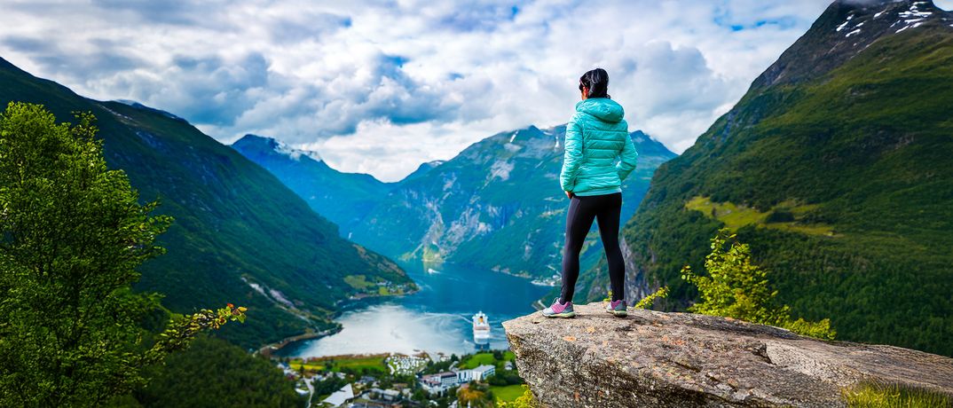 Eine Frau steht auf einem Felsvorsprung und schaut in einen Fjord hinunter wo ein Schiff zu sehen ist