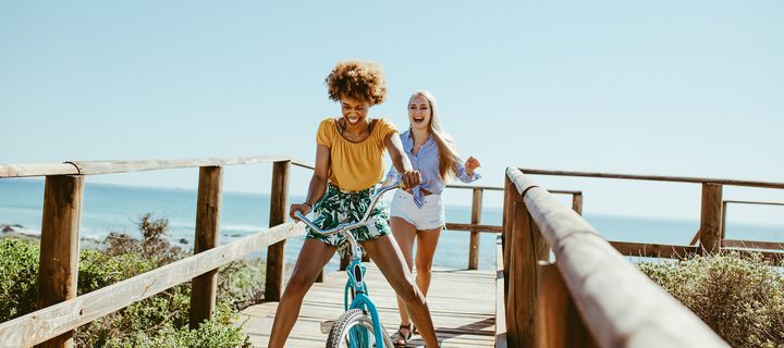 Frau, gefolgt von weiterer Frau, fährt mit Fahrrad über Holzsteg am Strand bei sonnigem Wetter