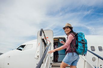 Eine Reisende mit Rucksack steigt über die Gangway in ein Flugzeug unter leicht bewölktem Himmel.