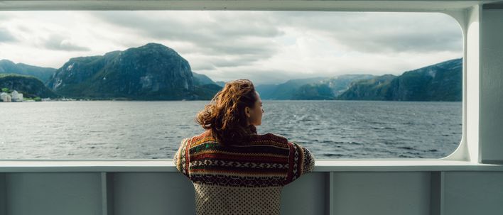 Eine Frau steht an der Reling eines Kreuzfahrtschiffs und blickt auf einen Fjord mit Bergen unter grauen Wolken.