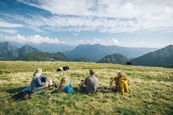 Wandergruppe genießt Rast auf grüner Almwiese mit Bergpanorama im Hintergrund