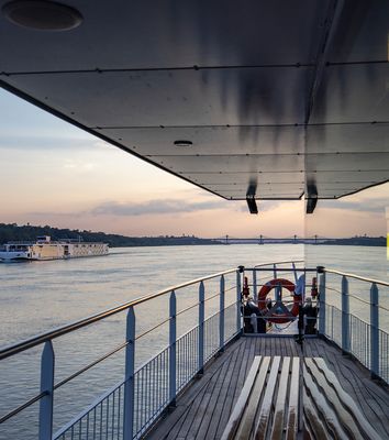 Blick auf das Außendeck eines Flusskreuzfahrtschiffes bei Sonnenuntergang mit Flusslandschaft im Hintergrund
