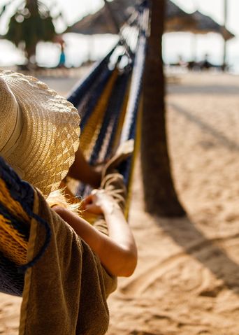 Person entspannt in einer Hängematte am sonnigen Sandstrand mit Strohhüten im Hintergrund