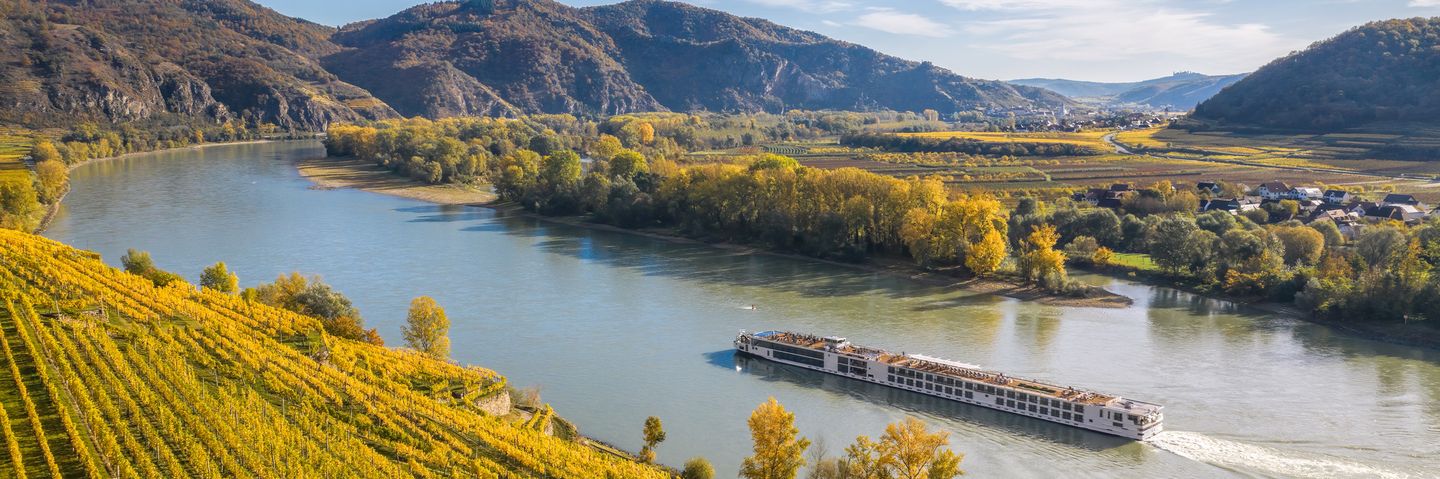 Flusskreuzfahrtschiff fährt entlang der Donau in Österreich mit Weinbergen am Ufer