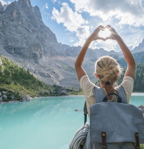 Frau von hinten fotografiert, steht vor einer weitläufigen Seenlandschaft in Italien und fängt mit ihrer zum Herz geformten Hand die Sonne ein
