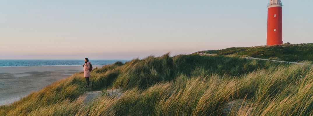 Frau steht in einer Dünenlandschaft vor einem Leuchtturm auf der Insel Texel