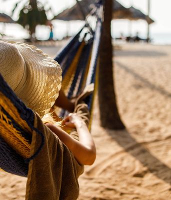 Person entspannt in einer Hängematte am sonnigen Sandstrand mit Strohhüten im Hintergrund