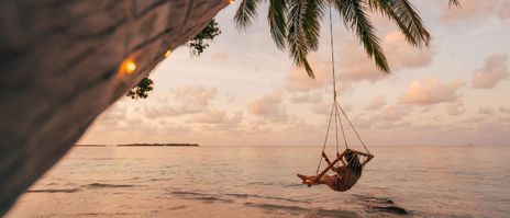 Frau in Hängeschaukel unter Palmen am Strand mit Blick aufs Meer im Abendlicht