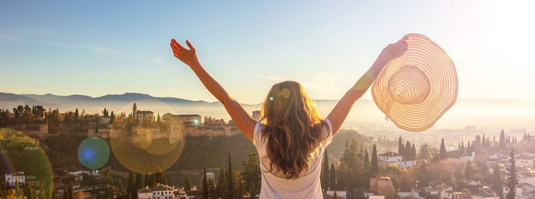Eine Frau breitet auf einem Aussichtspunkt über Granada die Arme aus und hält ihren Sonnenhut in die Höhe.