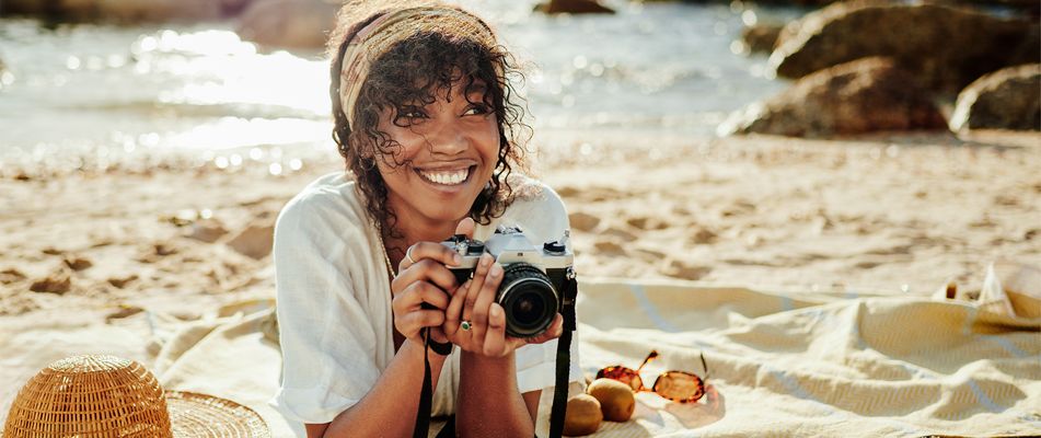 Frau mit Kopftuch und Kamera in der Hand liegt am Strand vor dem Meer mit Felsen in Sepiafarbton
