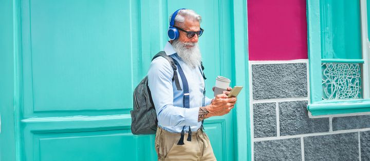 Mann mit grauem Bart, Kopfhörern und To-Go-Kaffee steht vor türkis-pinker Hauswand und guckt in sein Handy