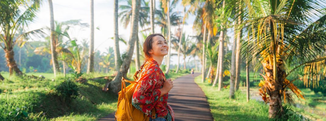 Reisende Frau mit Rucksack auf einem Weg zwischen Palmen in tropischer Landschaft auf Bali