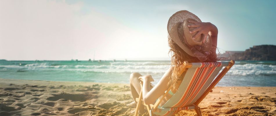 Frau sitzt im Sonnenliege am Strand mit Blick auf das Meer und hält ihren Sonnenhut fest