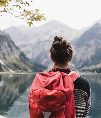 Blick auf die Rückansicht einer Frau mit Rucksack, die vor einem Bergsee auf die Berge blickt