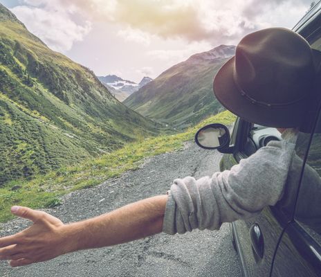Ein Reisender mit Hut und Rucksack öffnet die Arme vor grünen Bergen und einem weiten Tal.