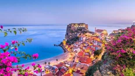 Panoramablick auf die Küste von Scilla in Kalabrien mit bunten Häusern am Hang, blauem Meer und pink blühenden Bougainvillea im Vordergrund