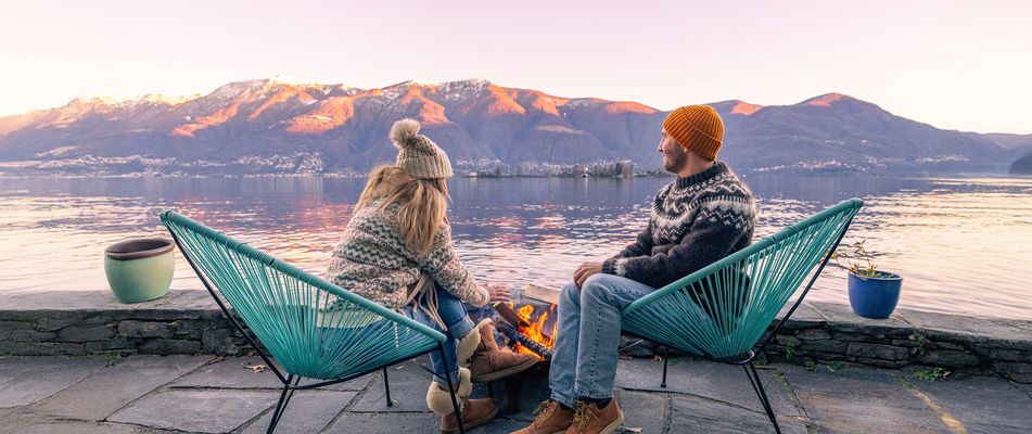 Paar sitzt am Lagerfeuer in Outdoor-Stühlen am Seeufer mit Bergpanorama bei Sonnenuntergang