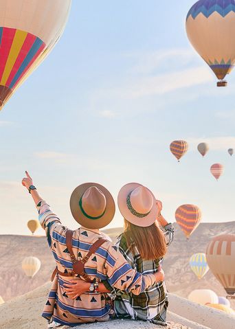 Pärchen in Rückansicht mit Hüten sitzt auf einem Felsen und zeigt auf die Heißluftballons am Himmel von Kappadokien in der Türkei