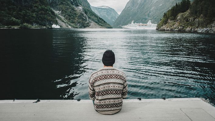 Ein Mann, der in einem Fjord auf einem Steg sitzt und auf ein Kreuzfahrtschiff in der Ferne schaut