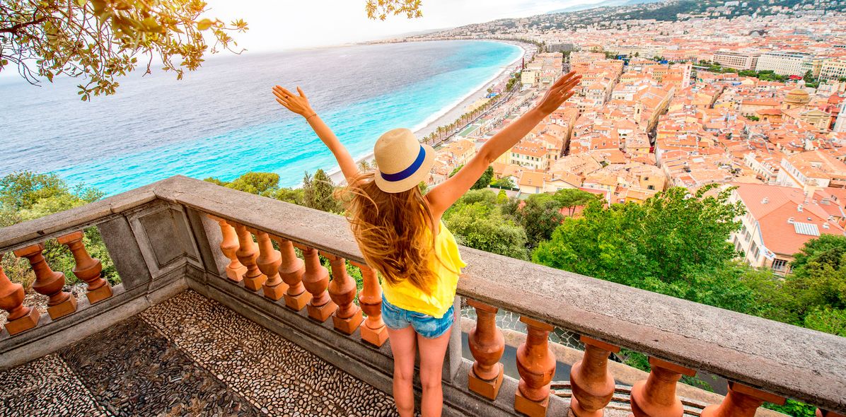 Frau mit Hut und ausgestreckten Armen blickt von einer Aussichtsterrasse über die Küste und Stadt von Nizza