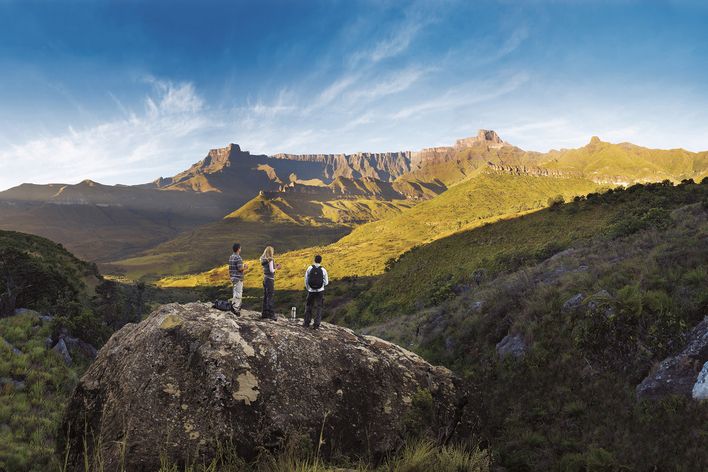 Drei Wanderer stehen auf einem felsigen Aussichtspunkt vor der weiten Bergkulisse der Drakensberge