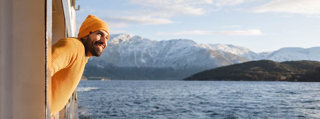 Ein Mann mit Mütze lehnt sich aus der Schiffstür und blickt über das Wasser auf schneebedeckte Berge.