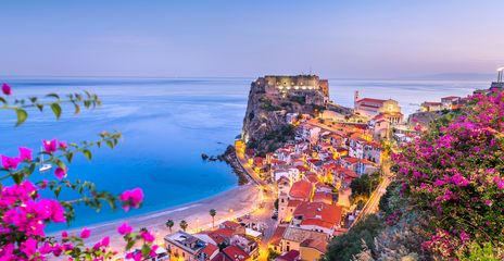 Panoramablick auf die Küste von Scilla in Kalabrien mit bunten Häusern am Hang, blauem Meer und pink blühenden Bougainvillea im Vordergrund