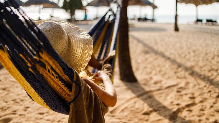 Person entspannt in einer Hängematte am sonnigen Sandstrand mit Strohhüten im Hintergrund