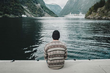 Ein Mann, der in einem Fjord auf einem Steg sitzt und auf ein Kreuzfahrtschiff in der Ferne schaut