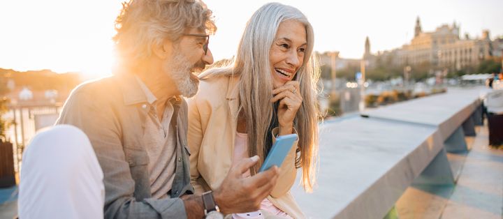 Best-Ager-Paar sitzt am Geländer, hält Kaffee in der Hand und lacht über etwas auf dem Smartphone