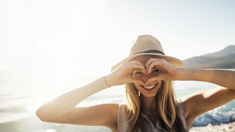 Frau mit Sommerhut am Strand formt mit den Händen ein Herz und schaut hindurch