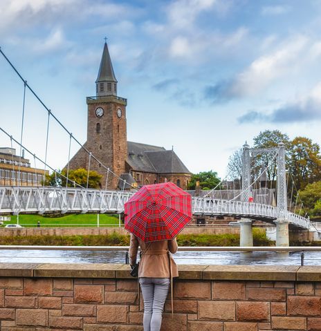 Frau mit rotem Regenschirm steht am Fluss Ness mit Blick auf die Brücke der Stadt Inverness