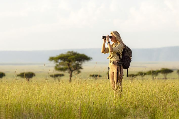 Frau steht während einer Safari mit Fernglas in weiter Graslandschaft mit Akazienbäumen im Hintergrund 