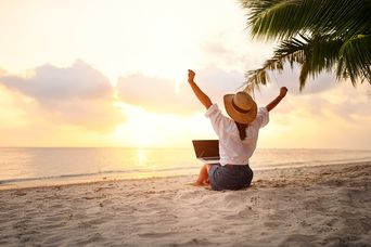 Frau sitzt mit Laptop am Strand vor dem Meer und genießt den Sonnenaufgang