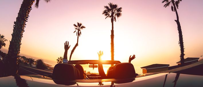 Zwei Frauen sitzen in einem Cabrio welches der Sonne entgegen und von Palmen umgeben auf einem Parkplatz steht und die Hände in die Höhe strecken