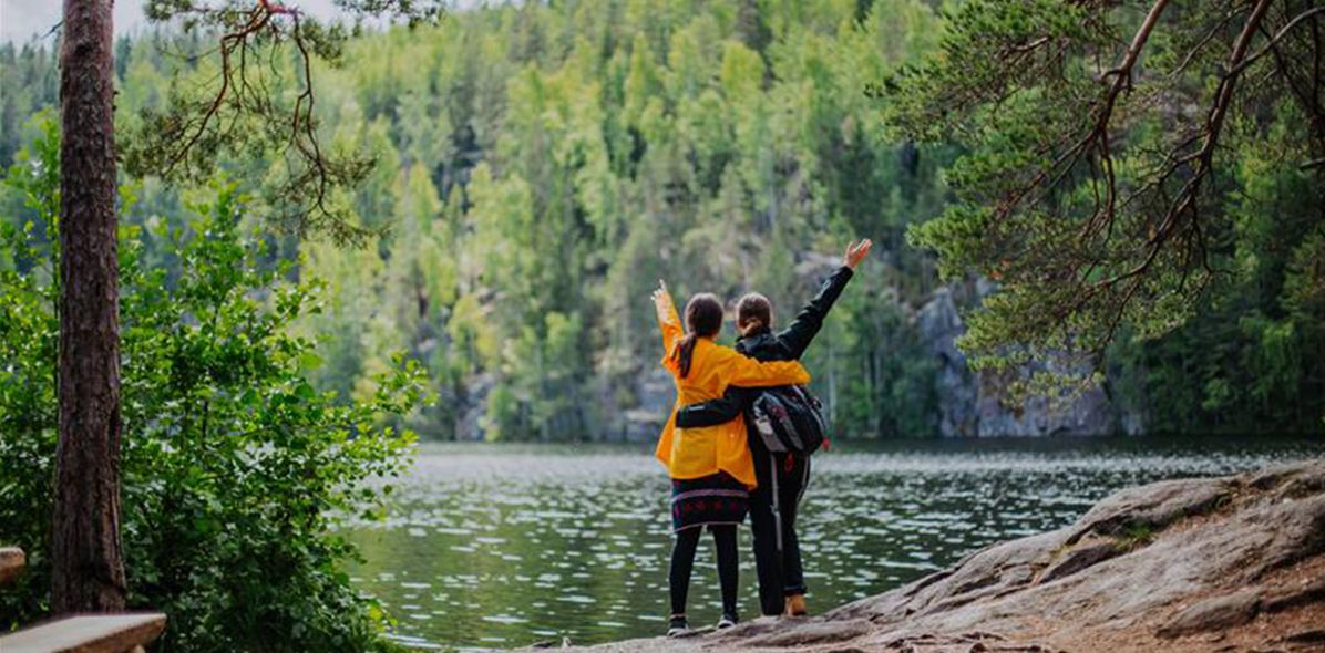 Zwei Reisende stehen am Seeufer in Jyväskylä, Finnland, und blicken auf die bewaldete Landschaft